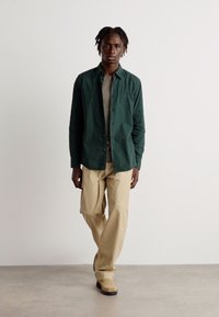 Camisa verde de botones con un corte relajado, superpuesta sobre un suéter beige claro, combinada con pantalones beige y botas marrones sobre un fondo neutro.