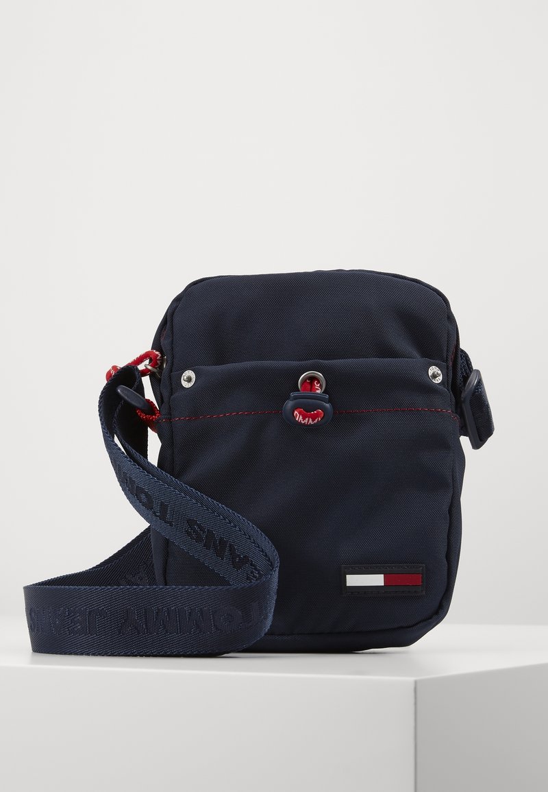 Borsa a tracolla blu realizzata in tessuto resistente, con dettagli rossi, un tirante con logo e una tracolla brandizzata con una patch rettangolare del logo.