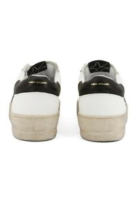 Un paio di sneakers bianche con accenti neri sul tallone e suole beige consumate viste da dietro, che mostrano il logo AMA-BRAND in lettere dorate.