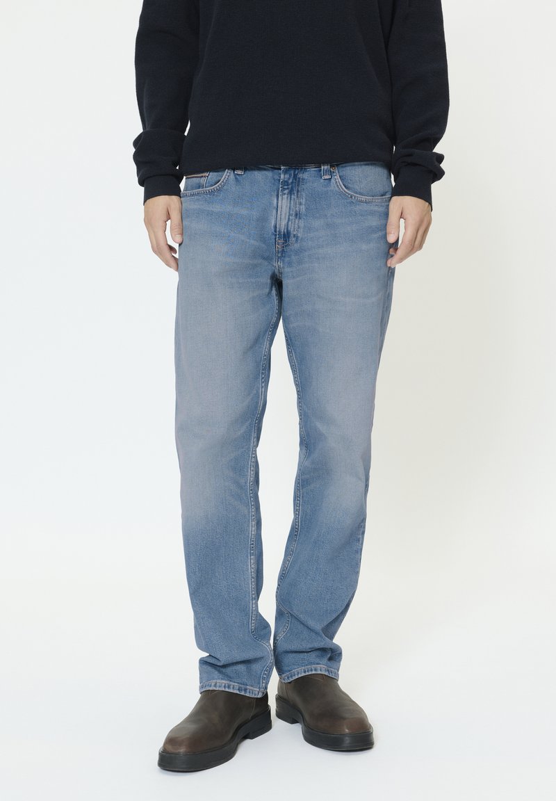 Moški, oblečen v svetlo modre kavbojke z ravnimi hlačnicami, črni pulover in temno rjavi usnjeni škornji, stoji na enobarvnem ozadju.