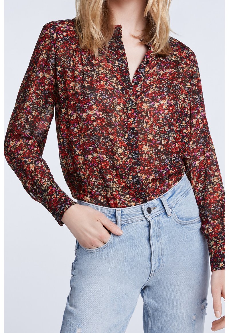 Blouse noire transparente avec un motif floral coloré, devant à boutons, manches longues et poignets froncés, associée à un jean taille haute bleu clair.