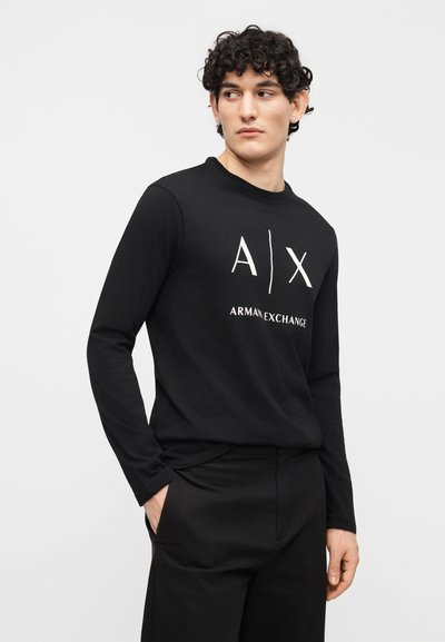 Млад мъж в черна риза с дълъг ръкав с лого "A|X Armani Exchange", черни панталони, стоящ с една ръка в джоба, гледащ настрани.