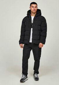 Schwarze Steppjacke mit Kapuze, gestepptem Design und Vorderreißverschlüssen. Über einem weißen Grafik-T-Shirt getragen, kombiniert mit schwarzen Cargo-Hosen und Sneakers.