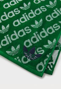 Zaļš dvielis ar iztīrītu "adidas" tekstu un trīszaru logotipa rakstu baltā un tumši zilā krāsā. Mīksta tekstūra ar kontrastējošām malām un izteiksmīgu dizainu.