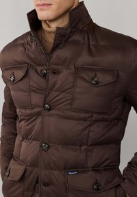 Chaqueta acolchada marrón con cuello alto, que presenta cuatro bolsillos delanteros, botones negros y un acabado texturizado. Tejido interior suave visible.
