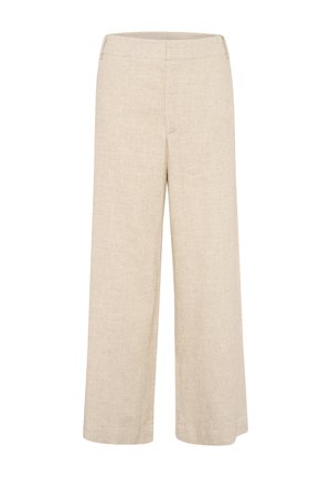 Femme aux longs cheveux bruns portant un haut blanc cintré, un pantalon beige à jambes larges et des chaussures marron, debout devant un fond gris uni.