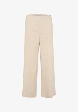 Pruunide pikkade juustega naine, kellel on seljas valge liibuv top, beežid laiad püksid ja pruunid kingad, seisab halli sirge tausta ees.