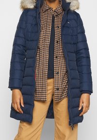 Marineblauwe pufferjas met een bontgevoerde capuchon, featuring een doorgestikte textuur, ritssluiting en zijzakken, gedragen over een bruine houndstooth shirt.