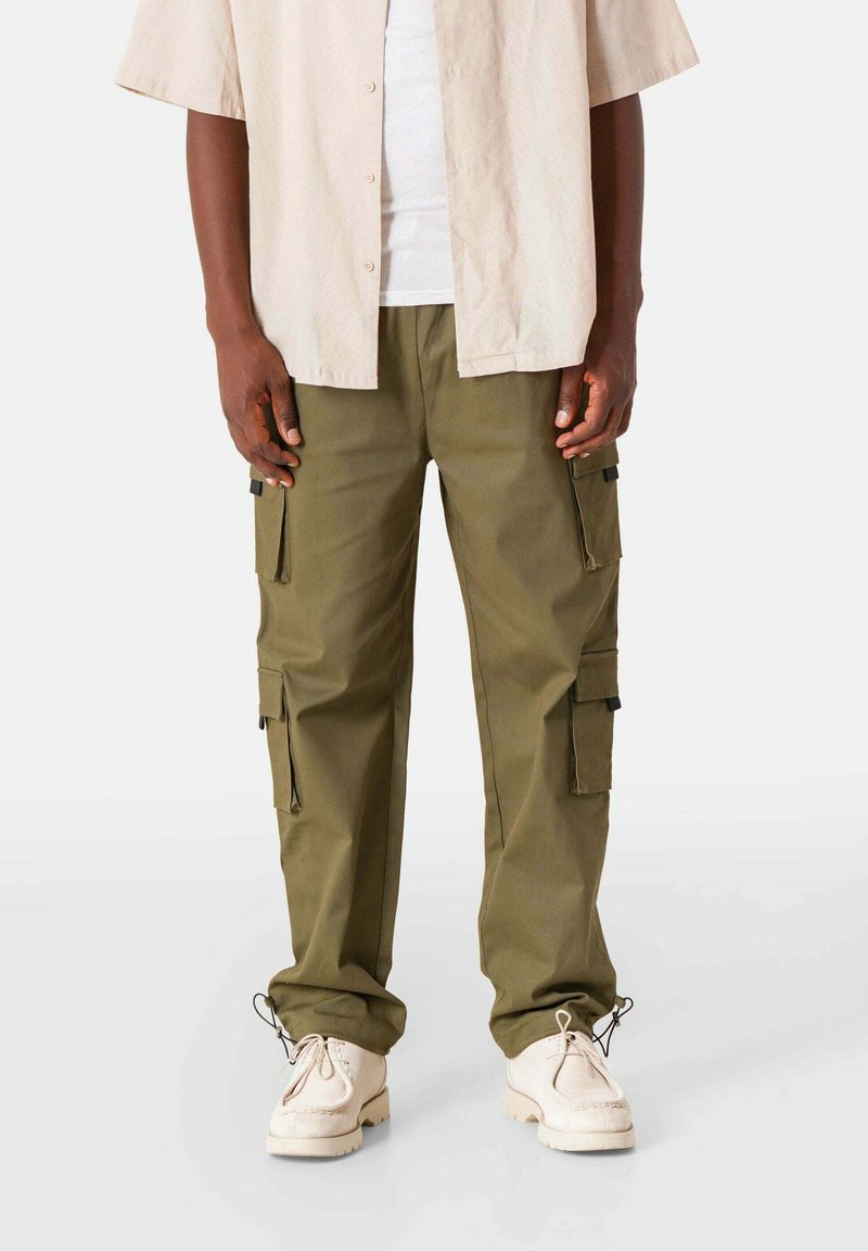 Olijfgroene cargobroek met meerdere zijzakken en een relaxte pasvorm, gecombineerd met beige schoenen en een licht knoopshirt.