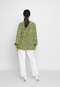 Černá košile s dlouhými rukávy a žlutým květinovým potiskem, volný střih a nařasené detaily na zadní straně, v kombinaci s bílými širokými kalhotami a podpatky.