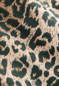 Leopardprintstof med mørkegrønne pletter på en beige baggrund. Teksturen er glat med et denimlignende udseende og synlige sømme.