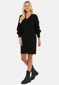 Schwarzes Strickkleid mit V-Ausschnitt und Zopfmuster, mit puffierten Ärmeln und lässiger Passform, kombiniert mit schwarzen Stiefeletten.