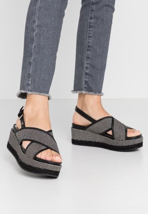 Pieds chaussés de sandales à plateforme rayées noir et blanc avec des brides croisées, associées à un jean gris à ourlet effiloché, sur un sol gris.