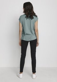 Teal t-shirt met korte mouwen en een ontspannen pasvorm, gecombineerd met zwarte slim-fit jeans en witte sneakers, vanuit de achterkant weergegeven tegen een witte achtergrond.