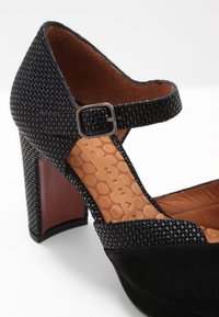 Chaussure à talons hauts noire avec une bande texturée ornée de motifs et une tige en daim. Dotée d'une sangle de cheville à boucle et d'une semelle intérieure marron.