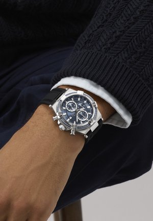Zápästie so striebornými chronografmi hodinkami, ktoré majú čierny ciferník, viacero subciferníkov a čierny gumový remienok, nosené cez čierny pletený sveter a manžetu bielej košele.