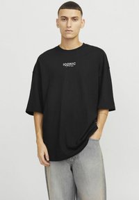 Czarny, oversize t-shirt z okrągłym dekoltem i krótkimi rękawami. Na klatce piersiowej znajduje się napis w białym kolorze "ICONIC". Noszony z jasnymi dżinsami.