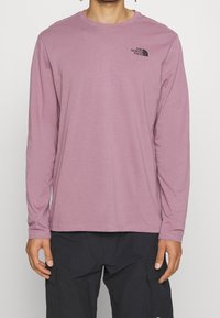 Světle růžové tričko s dlouhým rukávem vyrobené z měkké tkaniny, s černým logem na hrudi. Výstřih je kulatý a má rovné švy.