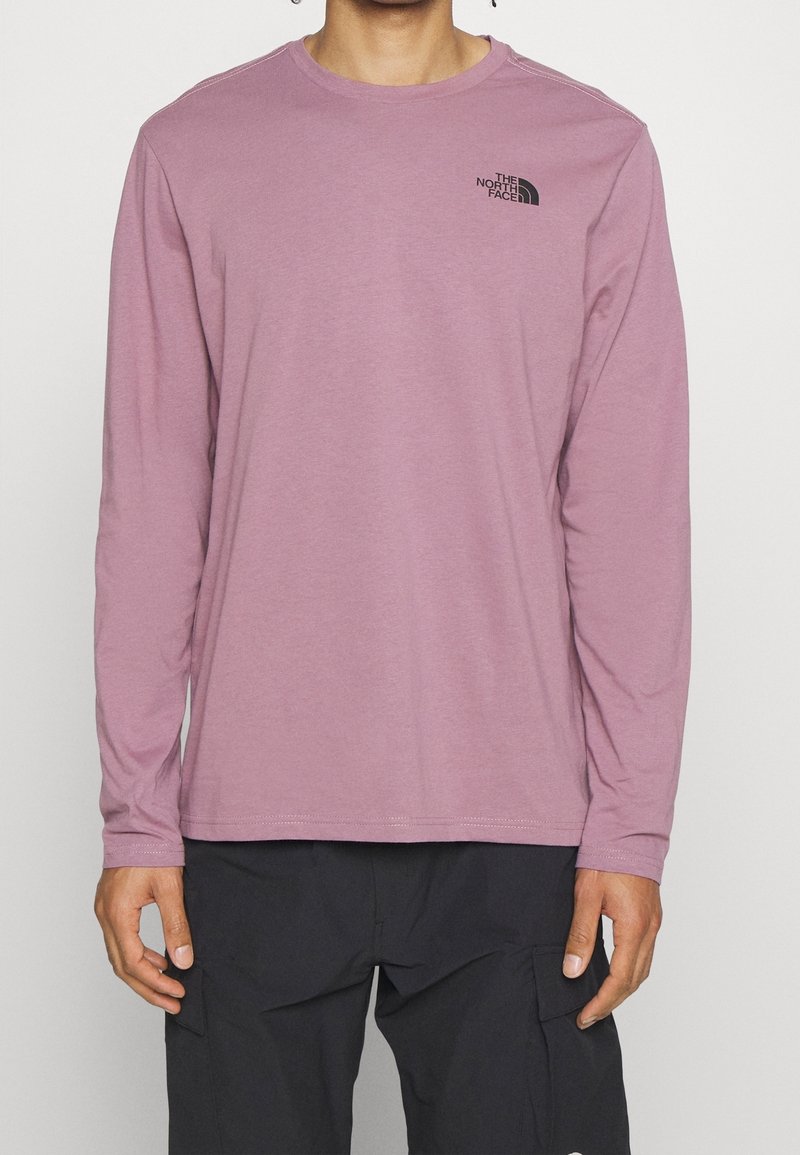 Světle růžové tričko s dlouhým rukávem vyrobené z měkké tkaniny, s černým logem na hrudi. Výstřih je kulatý a má rovné švy.