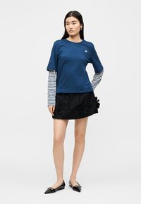 Γυναίκα με στρωματωτά μπλε T-shirts και μαύρη φούστα με ανθισμένες εφαρμογές, στέκεται μπροστά σε απλό φωτεινό φόντο.