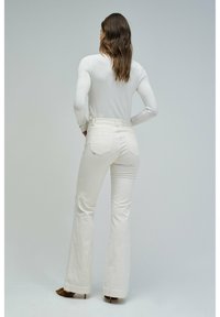 Pantalones acampanados blancos hechos de un tejido suave, con acentos cosidos y bolsillos traseros. Combinados con una blusa blanca de manga larga.