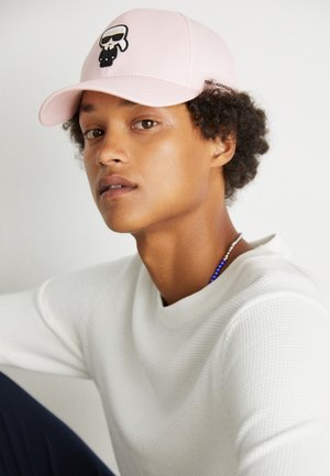 Ung person med krøllet hår iført en lyserød kasket med en tegneseriekranie og en hvid tekstureret sweater, der kigger let til siden.