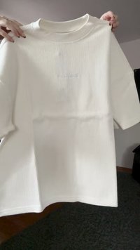 Hände halten ein einfaches, weißes, geripptes T-Shirt mit kleinen, schwachen Text auf der Brust vor einem neutralen Innenhintergrund.