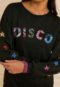 Fekete kapucnis pulóver, amelyen csillogó "DISCO" felirat található élénk színekben és csillagmotívumokkal piros, kék és rózsaszín színben az ujjakon. Texturált mandzsetták.