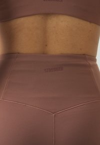 Brunt aktivtøy sett som inkluderer en tettsittende sports-bh og høytlivs tights. Mykt, elastisk materiale med "STRONGER" preget på midjebeltet.