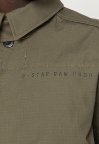 Camicia verde oliva con colletto strutturato, caratterizzata da un motivo a griglia, bottone a pressione e stampa con il marchio "G-STAR RAW PROD.".