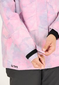 Leichte Jacke in pastellrosa und lila Tönen mit strukturiertem Muster, ausgestattet mit einem schwarzen Klettverschluss am Ärmel und einem Logo am Saum.