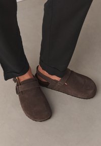 Personne portant des sabots en daim marron foncé à enfiler avec des brides réglables et un pantalon noir, debout sur un sol gris clair.