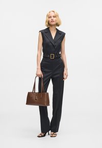 Čierny bezrukávový overal s priliehavým pásom a zlatou prackou, skombinovaný s hnedou koženou kabelkou a čiernymi sandálmi na vysokom podpätku.