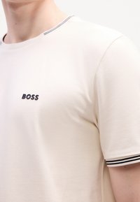 T-shirt beige à manches courtes en tissu texturé, avec un logo "BOSS" noir et des accents à rayures sur le col et les manches.
