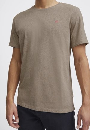 Mann trägt ein schlichtes hellbraunes kurzärmeliges T-Shirt mit einem kleinen roten bestickten Logo auf der linken Brust, kombiniert mit dunklen Hosen.