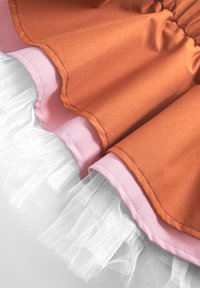 Oranssi satiinikangas vaaleanpunaisella reunuksella, kerrostettuna valkoisella tüllillä. Ominaisuuksiin kuuluu rypistynyt tekstuuri ja röyhelöhöyhen, joka luo tilavan ilmeen.