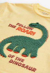 Gelbes T-Shirt mit einem grünen, strukturierten Dinosaurier-Graphic, roten Akzenten und gesticktem Text: "Folge dem BRULLEN! des DINOSAURIERS."