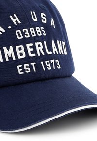 Sötétkék baseballsapka, amely szövetből készült, fehér hímzett felirattal: "TIMBERLAND EST 1973" és "N.H. USA 03885" az elején.