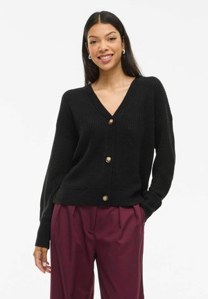Gilet noir à côtes avec un col en V, comportant six boutons dorés sur le devant. Manches longues et coupe décontractée, porté sur un pantalon bordeaux.