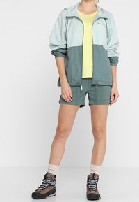 Ljusblå och grön vindtät jacka med elastiska ärmar, gul t-shirt, gröna shorts, beige vandringsstrumpor och bruna vandringskängor.