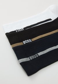 Tres calcetines doblados en blanco, negro con una franja marrón y azul marino con una franja plateada, cada uno con el logo "BOSS" cerca del borde.