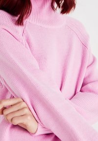 Pull rose en maille avec col roulé côtelé, manches longues et tissu texturé avec détails de coutures visibles.