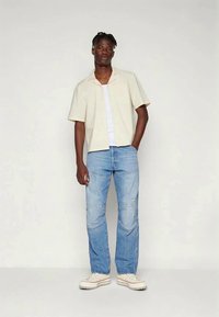 Κοντομάνικη μπεζ πουκαμίσα με κουμπιά, λευκό T-shirt από κάτω, ανοιχτό μπλε τζιν και μπεζ αθλητικά παπούτσια με υφασμάτινη σόλα και κόκκινες λεπτομέρειες.