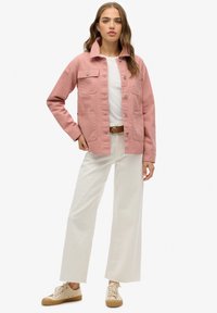 Giacca di jeans rosa con quattro tasche frontali, colletto e chiusura con bottoni. Abbinata a pantaloni bianchi a gamba larga e sneakers beige con lacci.