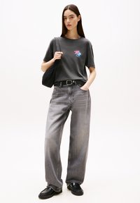 Camiseta de algodón gris con un gráfico en rosa y azul, combinada con unos vaqueros de denim gris sueltos. La modelo lleva un bolso tote negro y calza zapatos negros.