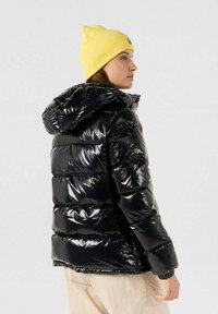 Giacca a vento nera con finitura lucida e trapuntata, dotata di cappuccio e polsini elasticizzati. Indossata con un berretto a maglia giallo.
