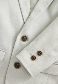 Blazer gris clair avec un tissu texturé, boutons marron et coutures détaillées sur les poignets pour une finition élégante.