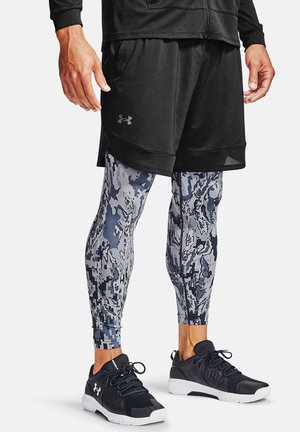 Man wearing black athletic shorts over gray and black camo-patterned compression leggings and black Under Armour sneakers with white soles.