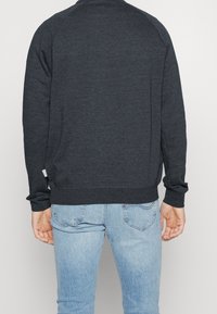 Sudadera de color gris oscuro con mangas largas, puños y dobladillo de canalé; textura suave; vista trasera sin gráficos ni patrones. Combinada con jeans de color azul claro.