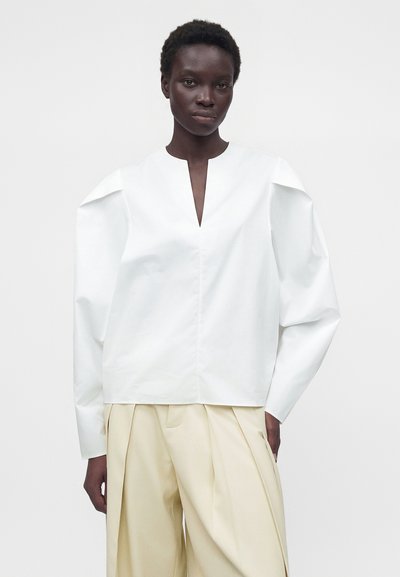 Femme portant un haut blanc à manches longues avec une fente en V et un pantalon beige large plissé sur un fond uni.